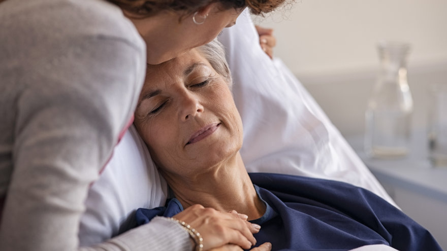 A moment of tenderness between a daughter visiting her mother recovering in the hospital. The mother is tunred towards her daughter, smiling with her eyes closed. The daughter is holding her hand while giving her a kiss on the forehead.