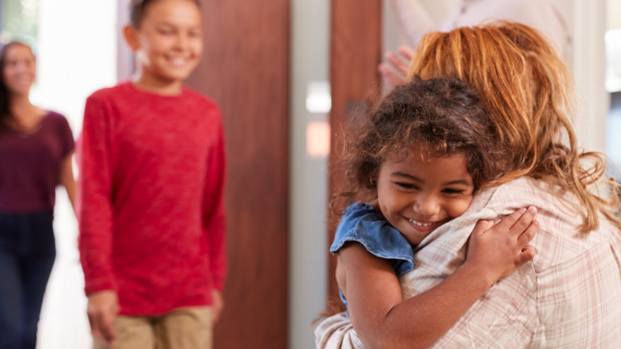 A young granddaughter being embraced by her grandmother while her brother and mother continue to walk into the house.