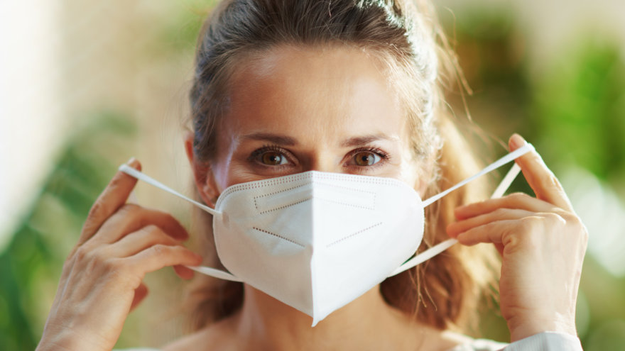 A smiling woman in the process of either putting on or taking off a face mask.